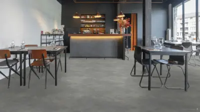 PVC-Boden Inspiration 4 Innenraum eines Restaurants mit grauem PVC-Boden in Betonoptik, mehreren gedeckten Tischen und einer Bar im Hintergrund. Große Fenster auf der rechten Seite sorgen für Tageslicht und betonen die gleichmäßige, moderne Bodenfläche.